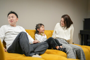 Wall Mural - A Japanese parent and child, a woman in her 30s, a man in his 20s, and a 6-year-old girl, are relaxing on a sofa in their living room.