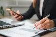 © Satori Studio - Close-up of a business professional reviewing financial charts and using a smartphone, showcasing modern office work.