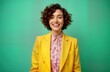 © Pete - Nonbinary person smiles brightly in studio portrait. Colorful casual attire featuring vibrant yellow blazer, pink floral shirt. Curly hair styled fashionably. Studio backdrop solid teal green.