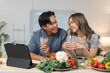 © crizzystudio - Young asian couple smiling and enjoying beverages while following a recipe on a tablet in their cozy kitchen, surrounded by fresh vegetables like cauliflower, bell pepper, and broccoli
