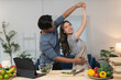 © crizzystudio - Young asian couple enjoying a lively dance while preparing fresh organic vegetables and fruits for a nutritious meal in their modern kitchen, radiating love and happiness