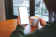 © Farknot Architect - Mockup image of a woman holding mobile phone with blank desktop screen while drinking coffee in cafe