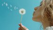 © DreamTone Creations - Dandelion dispersing wind. Young woman blowing a dandelion against a clear blue sky.