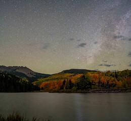 Wall Mural - Starry night sky over a serene mountain landscape with autumn foliage and a calm lake.