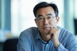 © STwul - Confident Asian businessman in glasses, hand on chin, thoughtful expression.