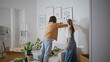 © stockbusters - Married couple hanging artwork on wall new home. Spouses decorating living room