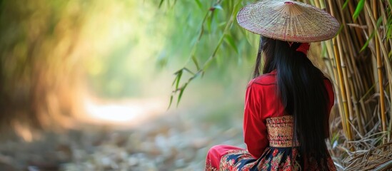 Elegant Asian woman in vibrant traditional costume sitting near bamboo grove in tranquil Thai countryside, reflecting and waiting, soft focus background