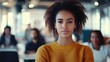 © 2rogan - Young black woman in a casual yellow sweater with an engaged expression leading a diverse group in a modern startup office with blurred colleagues in the background.