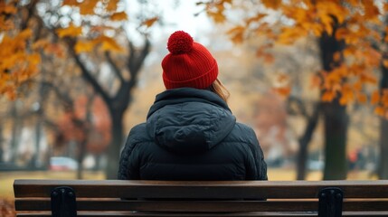 Wall Mural - Relaxing woman in red beanie and black jacket sitting on park bench surrounded by autumn foliage vibrant orange and yellow leaves mindful travel background