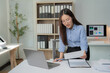 © Tj - Young Asian businesswoman standing at her desk reviewing documents and working on a laptop in a bright modern office, showcasing a productive and focused work environment