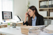 © Tj - Young businesswoman adjusting glasses and working with a pile of documents at the office desk, she is sitting at her desk and looking at the documents, she is wearing glasses and a black jacket