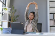 © amnaj - Young Asian businesswoman stretching her arms while sitting at her desk in a home office, taking a break from work