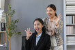 © amnaj - Two Asian businesswomen smiling and making an ok sign with their fingers, celebrating success and teamwork in a modern office setting