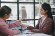 © amnaj - Two businesswomen exchanging a pen while engaging in a discussion during a meeting in a bright, modern office space