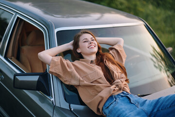 Canvas Print - Happy young woman relaxing on a vintage car, enjoying the outdoor sun, dressed in casual brown shirt and denim jeans, surrounded by green nature.