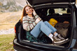 © maxbelchenko - Happy woman sits in the trunk of her car, parked on the side of a winding road that cuts through the rocky terrain. Lifestyle, travel, tourism, nature, active life