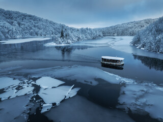 Wall Mural - Aerial drone view of boat in frozen lake at sunset. Winter fairytale in Plitvice Lakes, Croatia. Top view of boat, reflections in clear blue water, floating ice, snowy forest. White snow-covered trees