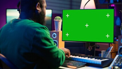 Poster - Musician operates on midi controller piano keys next to mockup screen, producing new songs with musical instruments and audio gear in home studio. Artist works with green screen. Camera A.