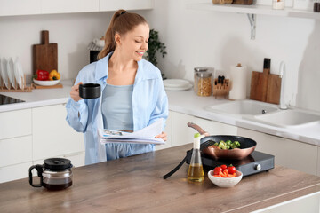 Wall Mural - Young woman with coffee and newspaper frying vegetables in kitchen