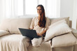 © SHOTPRIME STUDIO - Smiling woman sitting on a cozy sofa, typing on a laptop in a modern living room