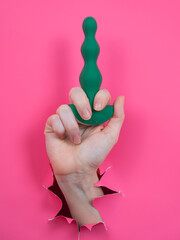 Wall Mural - Female hand sticking out with green anal beads from pink paper background. Vertical photo. 