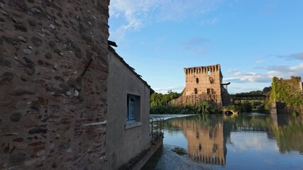 Wall Mural - Historic stone building by a calm river with an ancient tower and bridge in the background at sunset