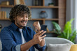 © Liubomir - Man with phone in hands celebrating victory and successful results of online achievement while sitting on sofa in living room at home in apartment.