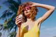 © SHOTPRIME STUDIO - Woman with curly hair enjoying ice cream under palm trees, wearing pink sunglasses and a yellow swimsuit, set against a bright blue sky