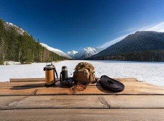 Poster - Winter camping gear rests on a wooden picnic table overlooking a snow-covered lake and majestic mountains under a vibrant blue sky.  A peaceful, scenic winter adventure awaits.