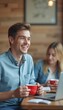 © natakot - A cheerful young man with light brown hair holds a red cup, radiating happiness in a warm café setting. Behind him, a woman with long blonde hair is engaged with her laptop, creating a relaxed and