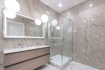 Stylish design of the bathroom decorated with beige marble tiles. Glass shower cabin. Stylish lamps.