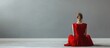 © 2rogan - Young woman in a vibrant red dress seated on light wooden floor against a muted gray wall, captured from a low angle for a minimalistic aesthetic.