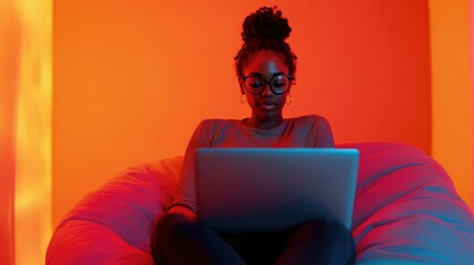 African American female programmer with eyeglasses using laptop on white beanbag chair in vibrant orange office lighting at night.
