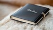 © Tati - Black leather-bound 'Manifest' notebook on plush beige surface