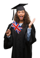 Wall Mural - Young hispanic woman wearing graduation uniform holding flag of UK very happy and excited, winner expression celebrating victory screaming with big smile and raised hands