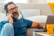 © simona - A 50-year-old man sits on the floor of his apartment, wearing casual clothes, talking on the phone to family or friends, enjoying his free time while relaxing in a quiet, comfortable home environment