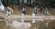 © ihorvsn - People actively collecting trash from ground. Volunteers working hard while trying keep nature clean. Pine forest in background. Trees mirroring from water surface. Wearing warm clothing.
