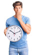 © Krakenimages.com - Young handsome man holding big clock covering mouth with hand, shocked and afraid for mistake. surprised expression