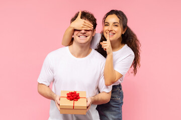 It's surprise. Lady covering her boyfriend eyes and making silence gesture, couple posing isolated over pink studio wall background