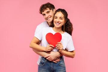 Young European couple in love embracing, holding paper heart, posing over pink studio background. Valentine's day celebration and romantic relationship