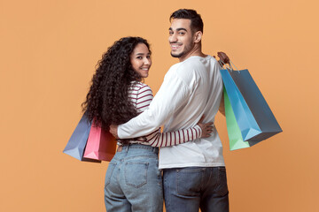 Family Shopping. Happy Arab Spouses Embracing And Holding Colorful Shopper Bags, Smiling Middle Eastern Couple Walking On Yellow Studio Background And Turning At Camera, Full Length, Copy Space