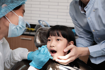 Wall Mural - Asian female pediatric dentist examines a girl's teeth in dental clinic and takes care and encourages of father, mouth oral hygiene, and professional orthodontic healthcare work in a kid hospital.