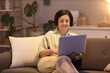 © Pixel-Shot - Mature woman using laptop on sofa at home in evening