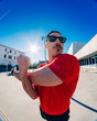 © Studio Marmellata - man doing sport in a red t-shirt and black shorts performs arm stretches under bright sunlight in a modern urban plaza with clean architectural lines and clear blue skies in the background
