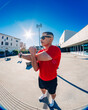 © Studio Marmellata - man doing sport in a red t-shirt and black shorts performs arm stretches under bright sunlight in a modern urban plaza with clean architectural lines and clear blue skies in the background
