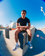© Studio Marmellata - A young man with short curly hair sits on a concrete bench in an urban outdoor space under a bright blue sky, wearing a black t-shirt, yellow shorts, and white sneakers, with a relaxed posture