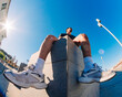 © Studio Marmellata - A young man in a black shirt and yellow shorts sits on a concrete urban structure in bright sunlight, captured with a fisheye lens against a clear blue sky and open urban space