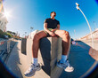 © Studio Marmellata - A young man in a black shirt and yellow shorts sits on a concrete urban structure in bright sunlight, captured with a fisheye lens against a clear blue sky and open urban space