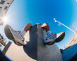 © Studio Marmellata - A fisheye lens close-up showing a young man's sneakers resting on a concrete ledge in an outdoor urban area with bright sunlight and clear blue skies in the background