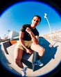 © Studio Marmellata - A young man in yellow shorts and white sneakers poses confidently on a concrete urban ledge captured under bright sunlight and a vivid blue sky using a fisheye lens perspective